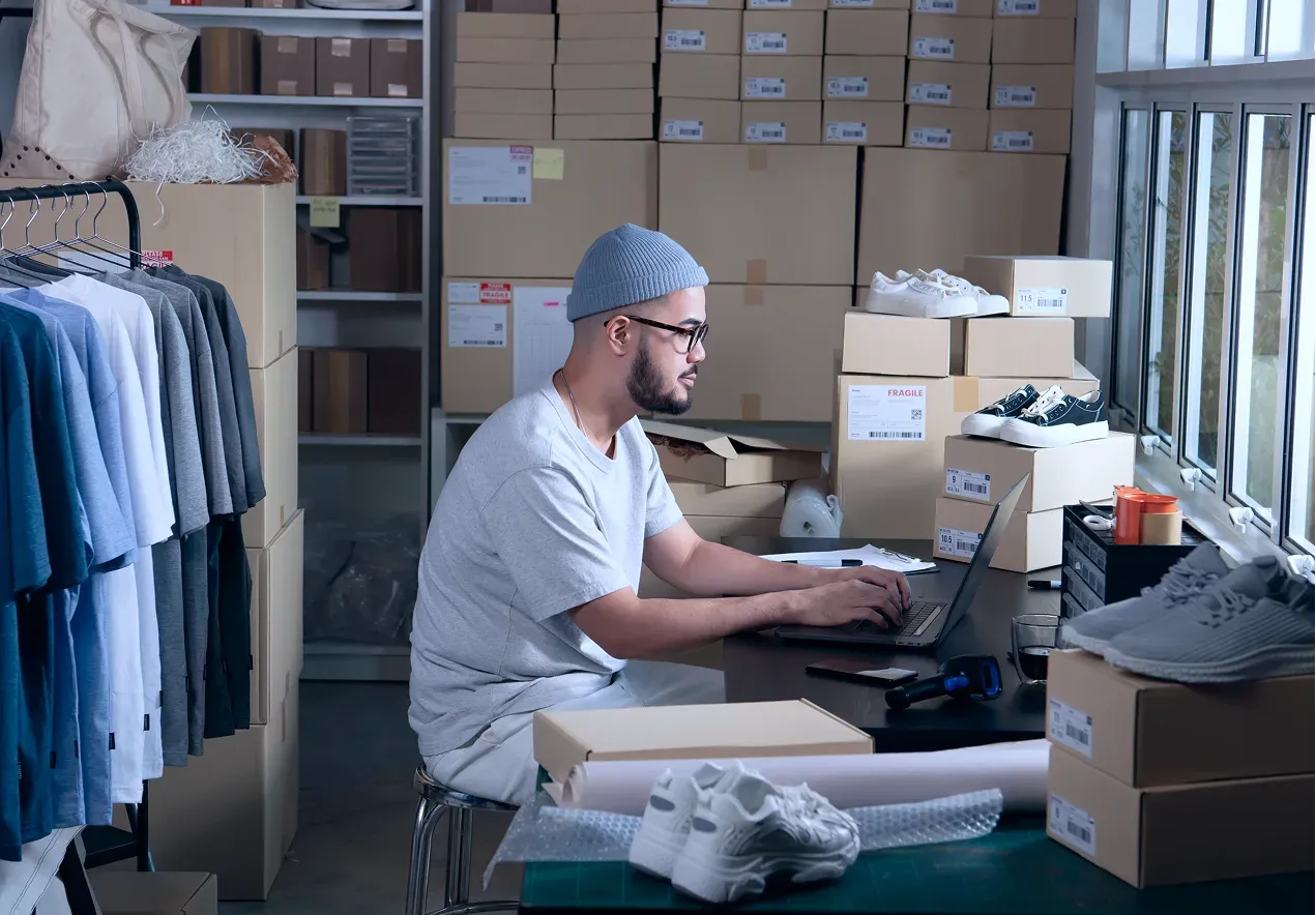 A small business owner working at a laptop among clothing racks and shipping boxes.